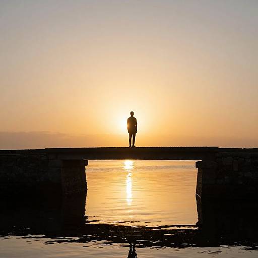 Photograph of a silhouetted person standing on a wooden pier at sunset, with the sun reflecting on calm water below.
