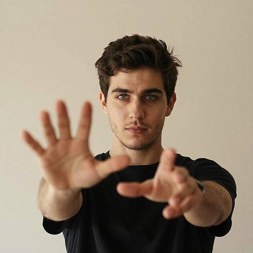 Photograph of a young, white man with short dark hair, blue eyes, and stubble, extending his hands forward, fingers spread, against a