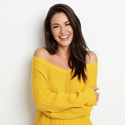 Photograph of a smiling brunette woman with long hair, wearing an off-shoulder yellow knit sweater, arms crossed, against a plain white background.
