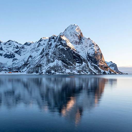 Photograph of a snow-capped mountain reflected in a calm, blue water body under a clear, light blue sky.