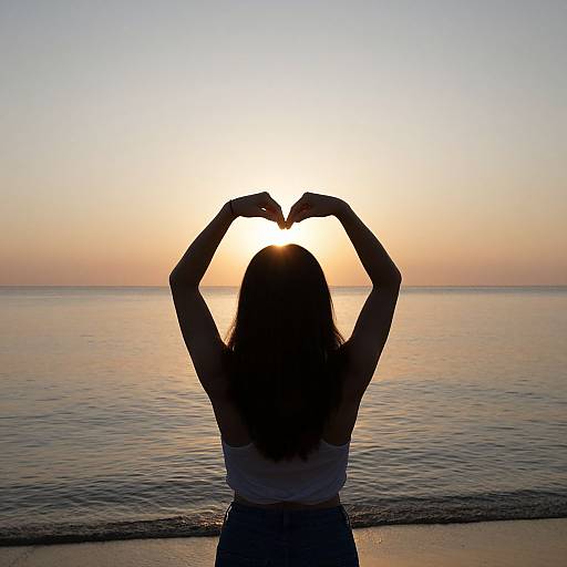 Photograph of a woman with long hair, silhouetted against a sunset, forming a heart shape with her hands, standing by a calm ocean