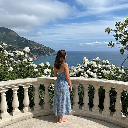 Photograph of a woman in a blue, patterned sundress, standing on a stone balcony, overlooking a coastal landscape with white flowers and a blue