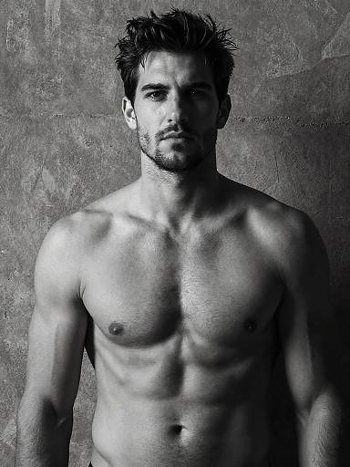 Black-and-white photograph of a muscular, shirtless man with short, spiked hair and a serious expression, standing against a textured gray background.