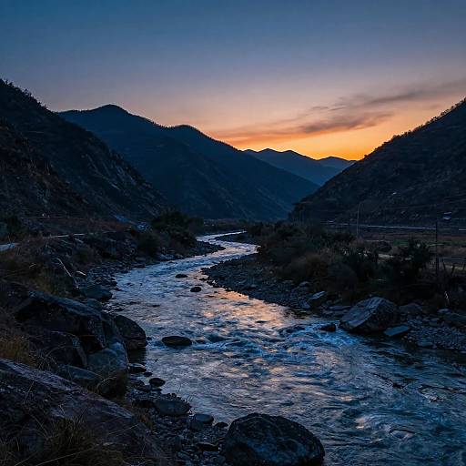 Photograph of a serene river flowing through a rocky valley at sunset, with dark silhouetted mountains and a vibrant orange-yellow sky.