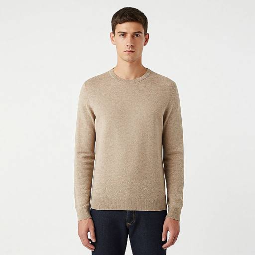 Photograph of a young man with short dark hair, wearing a beige knit sweater and dark blue jeans, standing against a plain white background.