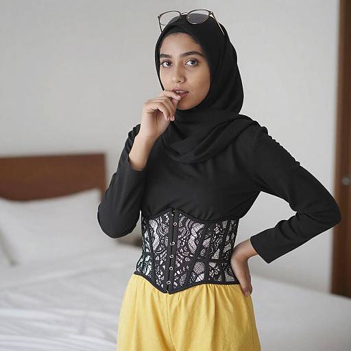 Photograph of a young woman with medium brown skin, wearing a black hijab, black lace corset, and yellow skirt, standing in a bright