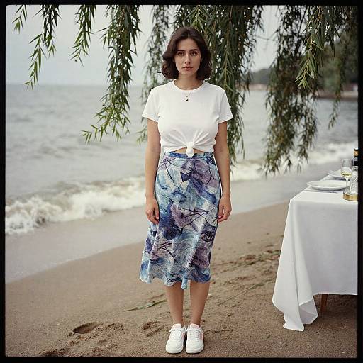 Young woman with shoulder-length dark hair, wearing a tied white top and blue floral skirt, stands on a sandy beach under leafy branches, with a