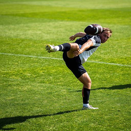 Photograph of a male rugby player in mid-action, wearing black and white uniform, holding a ball, on bright green grass field.