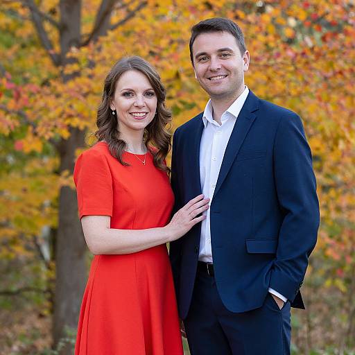 Photograph of a smiling Caucasian couple in autumn; woman in bright red dress, man in black suit and white shirt, standing close together. Background features