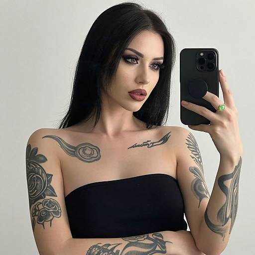 Tattooed woman with long black hair, dark red lipstick, black strapless top, taking a mirror selfie with a black phone.