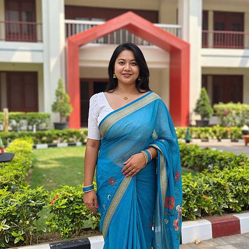 Photograph of an Indian woman with medium brown skin, black hair, wearing a blue saree with gold and red embroidery, white blouse, standing in