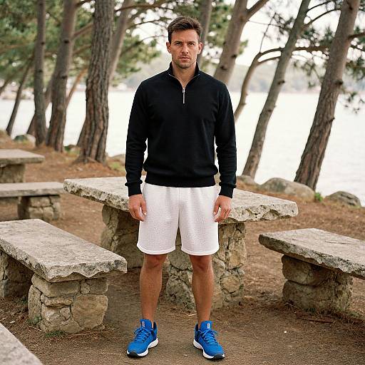 Photograph of a fit, Caucasian man with short brown hair, wearing a black zip-up hoodie, white shorts, and blue sneakers, standing in a