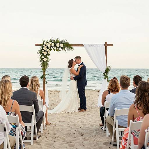 Photograph of a beach wedding: bride in white gown, groom in navy suit, kissing under floral arch, guests seated in white chairs.