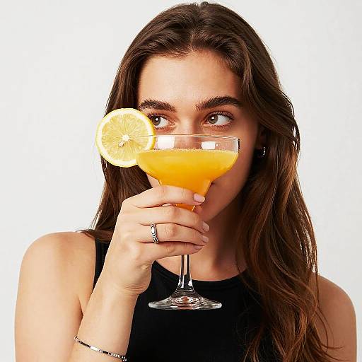 Photograph of a young woman with long brown hair, wearing a black sleeveless top, holding a glass of orange cocktail with a lemon slice, against