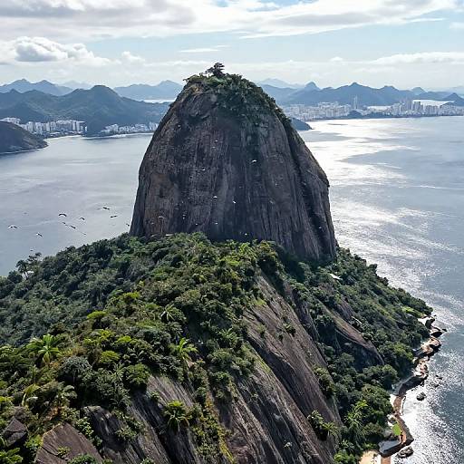 Aerial photograph of Sugarloaf Mountain in Rio de Janeiro, Brazil, with lush green foliage, sparkling ocean, city skyline, and cloudy sky.