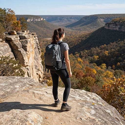 Photograph of a woman with dark hair in a ponytail, wearing black pants and a gray backpack, standing on a rocky cliff, facing a vast