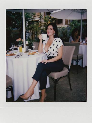 Photograph of a woman with dark hair, wearing a white polka-dot blouse and black pants, sitting at an outdoor café table, sipping coffee
