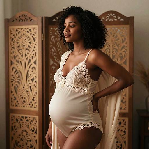 Photograph of a pregnant Black woman with curly hair, wearing a white lace-trimmed nightgown, standing before an ornate wooden folding screen