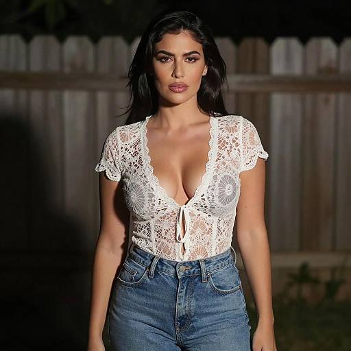 Photograph of a dark-haired woman with tan skin, wearing a deep V-neck white lace top and high-waisted blue jeans, standing in front