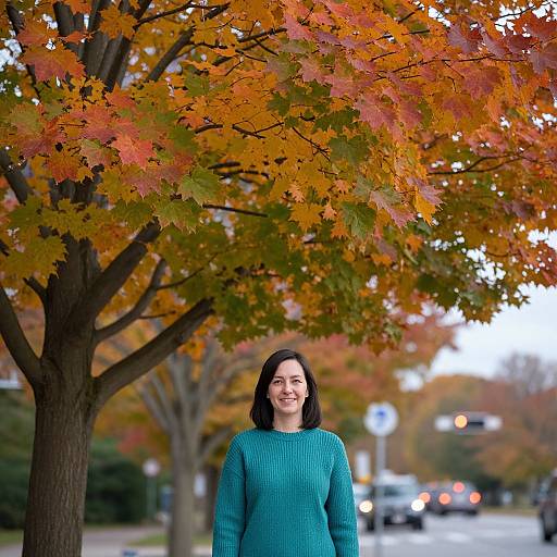 Photograph of a smiling woman with dark brown hair, wearing a teal sweater, standing under a tree with vibrant autumn leaves. Background includes blurred street and