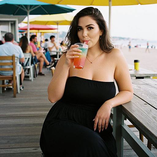 Photograph of a curvy woman with dark hair, wearing a black strapless dress, sipping a colorful drink at a beachside boardwalk.