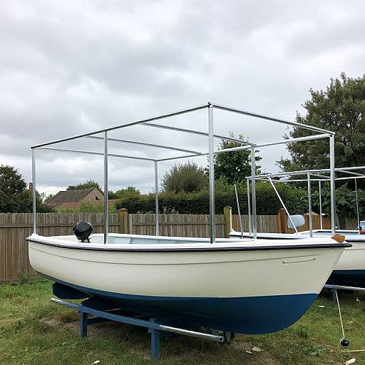 Photograph of a white and blue, small, boat with a metal frame canopy, on a grassy yard, with a wooden fence and trees in