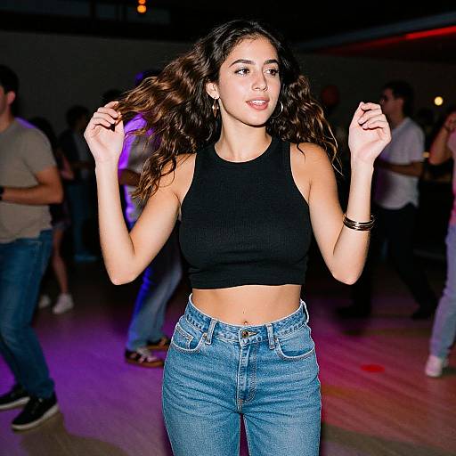 Photograph of a young woman with wavy brown hair, wearing a black sleeveless crop top and blue high-waisted jeans, dancing in a
