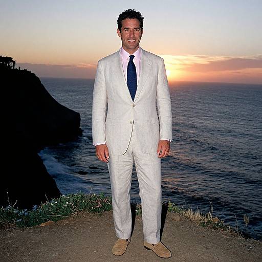 Photograph of a smiling man in a white suit, navy tie, and beige shoes standing on a cliff at sunset with a colorful sky and ocean in