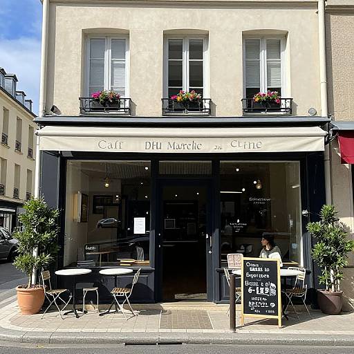 Photograph of a charming French café with beige facade, black-framed windows, flower boxes, outdoor tables, and a chalkboard menu.