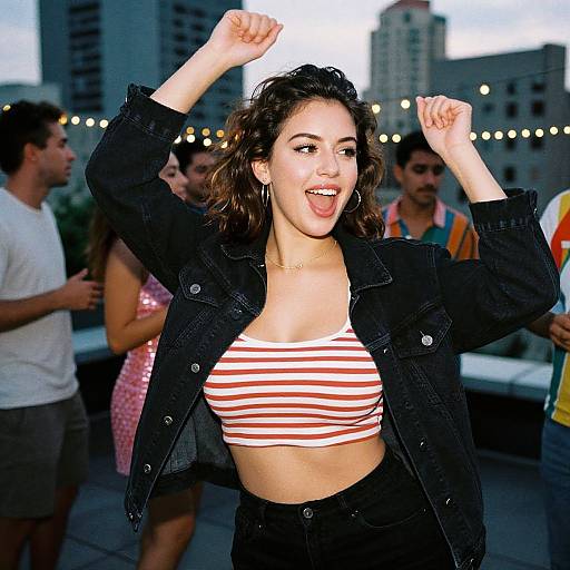 Photograph of a young woman with curly brown hair, wearing a black jacket and red-striped crop top, dancing with arms raised, surrounded by people in