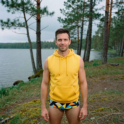Photograph of a muscular man with short brown hair, wearing a yellow sleeveless hoodie and colorful geometric-patterned briefs, standing by a lake with