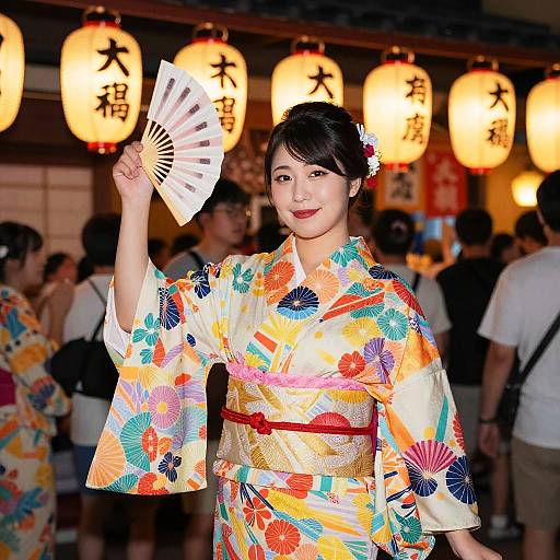 Japanese woman in colorful floral kimono holding fan, smiling, standing in front of glowing paper lanterns at night festival. Photograph.