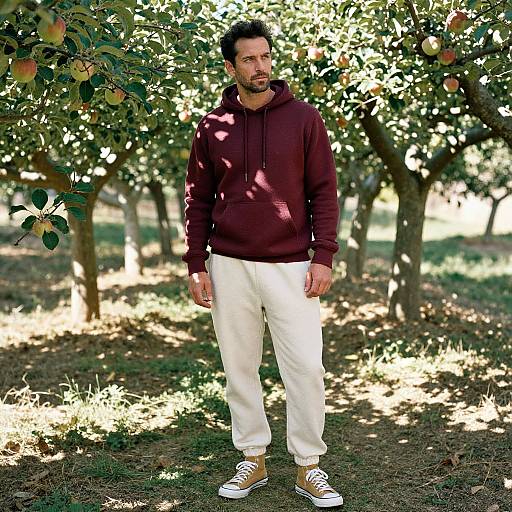 Photograph of a bearded man with dark hair, wearing a maroon hoodie, white pants, and yellow sneakers, standing in a sunlit apple