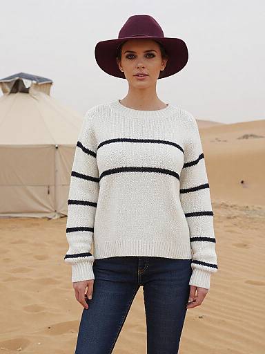 Photograph of a woman with fair skin, dark eyes, wearing a maroon hat, white knit sweater with black stripes, and dark jeans, standing
