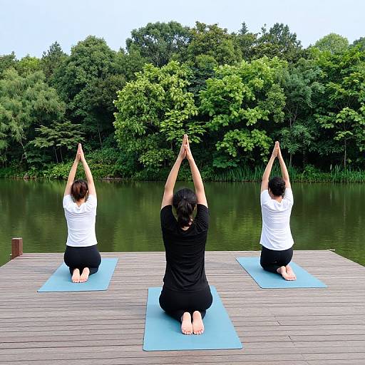 Photograph of three women in white shirts and black pants, kneeling on blue yoga mats on a wooden dock, arms raised, facing a lush green forest