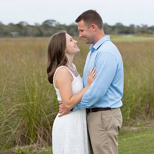 Photograph of a smiling couple standing in a grassy field, woman in white lace dress, man in blue shirt and beige pants, embracing lovingly