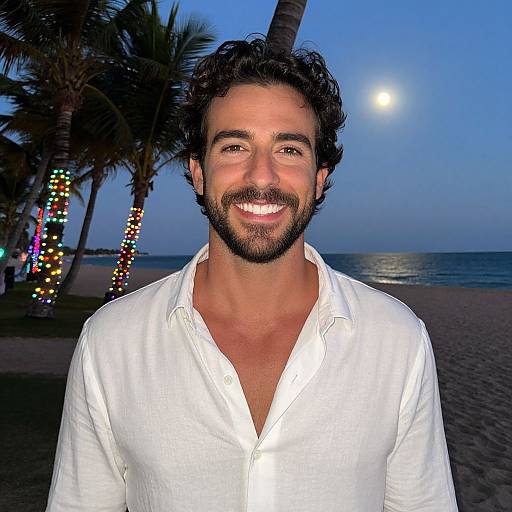 Photograph of a smiling, bearded man with dark curly hair, wearing a white button-up shirt, standing on a tropical beach at dusk with palm