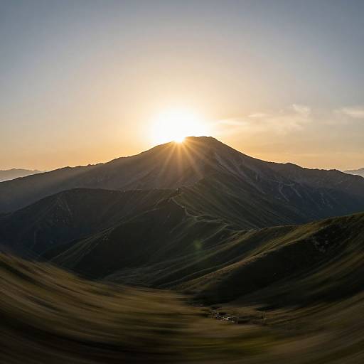 Photograph of a sunsetting mountain peak, casting radiant sunrays over dark, rolling hills with a small cluster of buildings in the foreground.