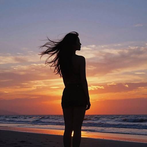 Silhouetted woman with long hair stands on beach at sunset, waves in foreground, vibrant orange and purple sky in background. Photograph.