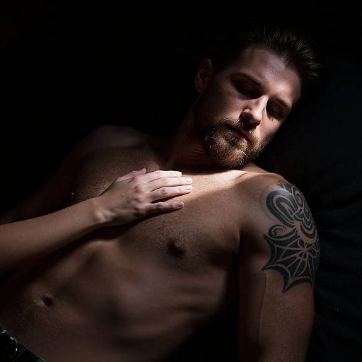 Photograph of a shirtless, tattooed man with dark hair, hand on chest, in low-light, shadowed background, emphasizing his muscular physique
