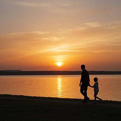 Silhouetted father and child holding hands walk by a serene lake at sunset, with a vibrant orange and yellow sky.