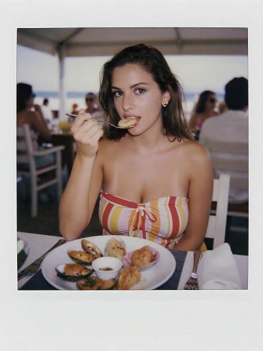 Photograph of a young woman with light skin and brown hair, wearing a strapless, striped top, eating a cookie at an outdoor beachside restaurant