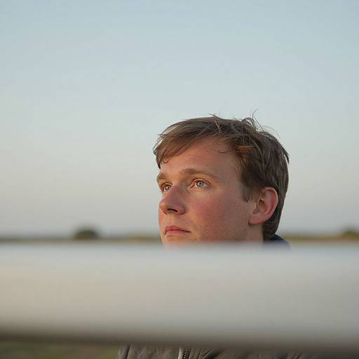 Photograph of a young white man with short brown hair and light blue eyes, gazing into the distance, blurred white railing in foreground, clear sky