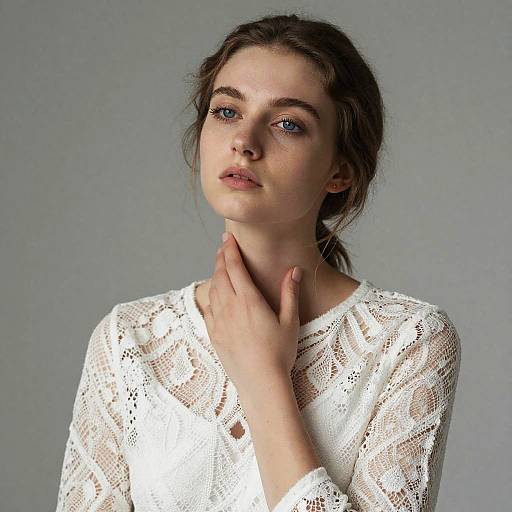 Photograph of a young woman with fair skin, blue eyes, and dark brown hair in a loose ponytail, wearing a white lace top, against