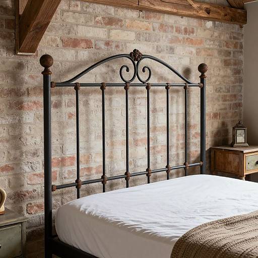 Photograph of a rustic bedroom with a black wrought-iron bed, white bedsheet, wooden nightstand, lantern, exposed brick wall, and wooden