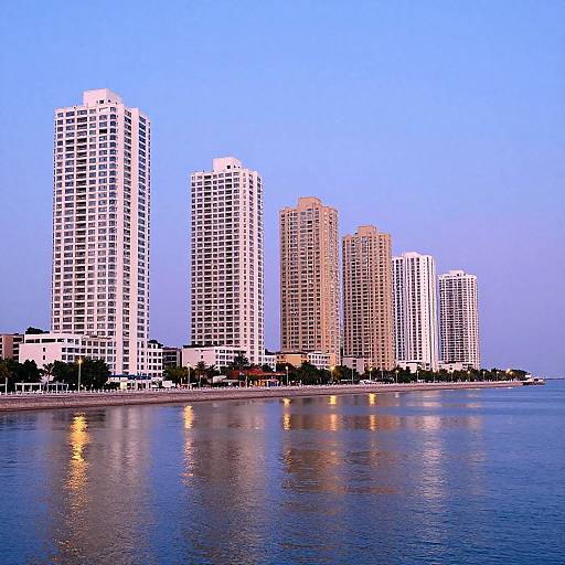 Photograph of tall, white and beige skyscrapers reflecting on calm water at dusk, with a clear blue sky background.