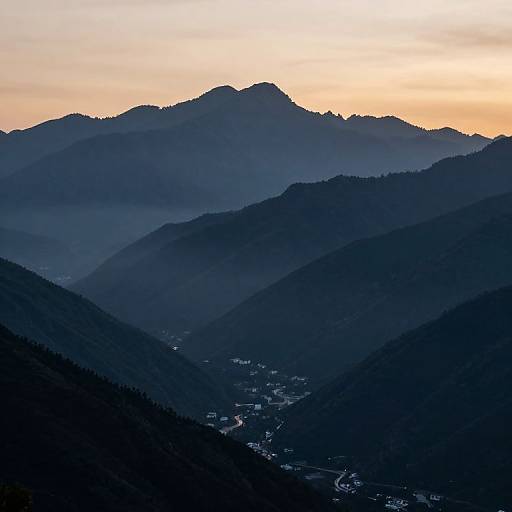 Photograph of a serene mountain landscape at sunset, showcasing layered silhouettes of dark blue mountains against a soft orange and yellow sky, with a small