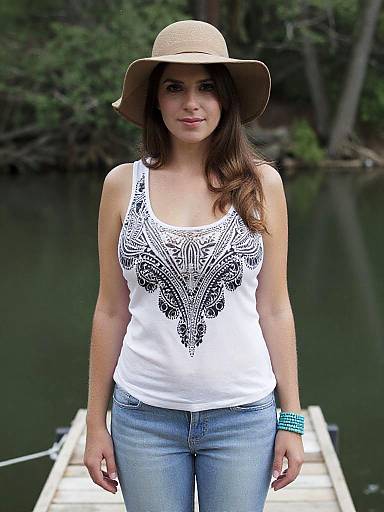 Photograph of a young woman with long brown hair, wearing a beige sun hat, white tank top with black mandala design, blue jeans, and