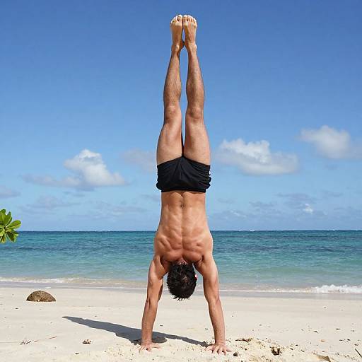 Photograph of a muscular, tan-skinned man with short black hair doing a handstand on a sunny beach, wearing black swim trunks, with