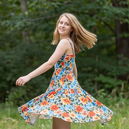Photograph of a smiling young woman with light brown hair in a colorful floral dress, dancing outdoors in a green forest.